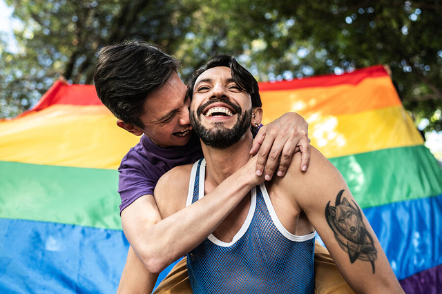 Gay couple piggyback outdoors with rainbow flag on background by Frazao Studio Latino